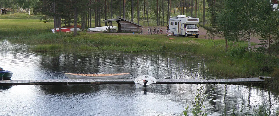 Stilla sjö med en brygga i förgrunden och en husbil och en liten stuga under höga tallar på andra stranden